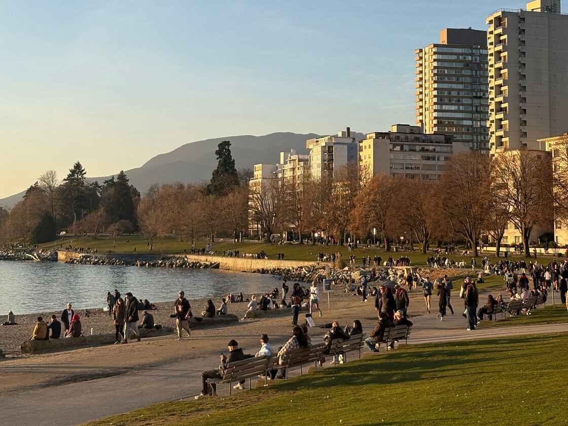 People sitting on benches, and gathered along a city beach, and seawall in late afternoon light, with apartment towers, and mountains behind them.