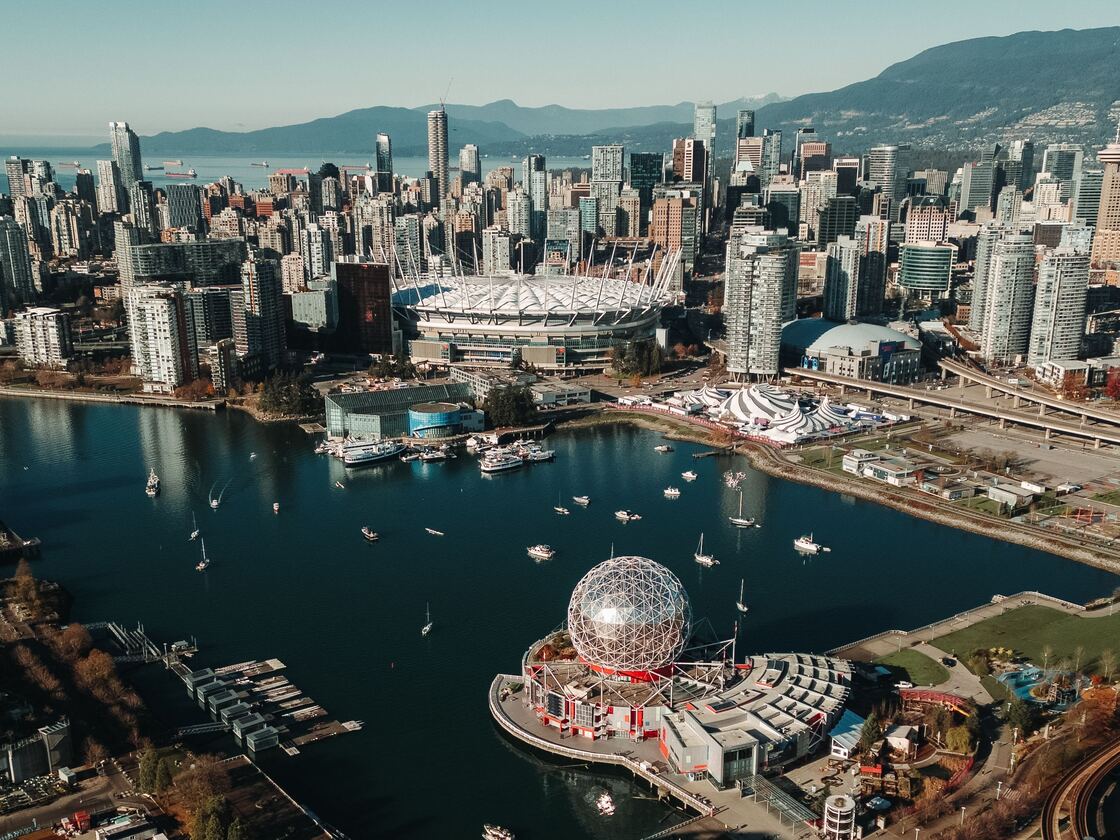 Aerial view over False Creek: Science World's geodesic dome, and the boats on the water in the foreground, BC Place stadium, and the downtown skyline behind, mountains beyond.
